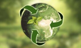 Rendering di un’immagine che rappresenta, su uno sfondo sfocato in cui si intravedono delle foglie e delle piante, la Terra, tutta colorata di verde a richiamare il concetto di ecologia. Ad avvolgere la Terra ci sono le tre frecce simbolo del “riciclo”